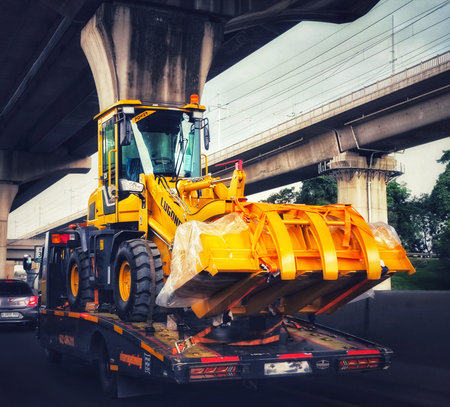 Cikarang, Indonesia on 30 Oct 2025. Bright yellow loader is transported on a black flatbed truck driving under a concrete overpass, conveying a sense of industry and transportation, perfect for construction or transport-related marketing.のeditorial素材