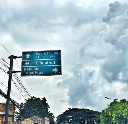 Bogor, Indonesia on 25 Oct 2025. Teal road sign displays directional information against an overcast sky, with verdant treetops visible at the bottom, creating a travel and destination themed composition suitable for tourism materials.のeditorial素材