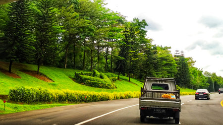 Bogor, Indonesia on 25 Oct 2025. Small white truck driving on asphalt road with green landscaped trees and grassy slopes under cloudy sky creates natural and calm scene suitable for travel or transportation themes.のeditorial素材