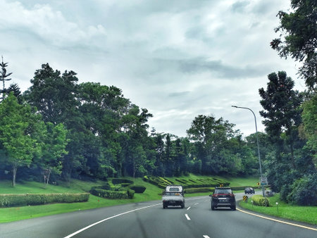 Bogor, Indonesia on 25 Oct 2025. Vehicles drive along a wet, gray highway lined with lush green trees and sculpted hedges under an overcast sky, conveying a sense of travel and outdoor scenery suitable for travel and lifestyle content.のeditorial素材
