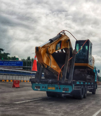 Cikampek, Indonesia on 20 nov 2025. Yellow excavator secured on a flatbed truck is being transported along a highway under a cloudy sky, ideal for industrial or construction themes.のeditorial素材