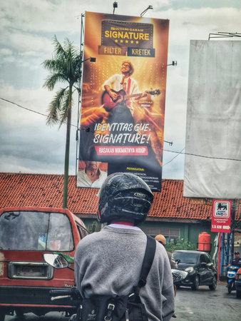 Bekasi, Indonesia on 25 Sep 2025. A motorcyclist wearing helmet and gray jacket looks up at a tall city billboard advertising music or event on a busy street.のeditorial素材