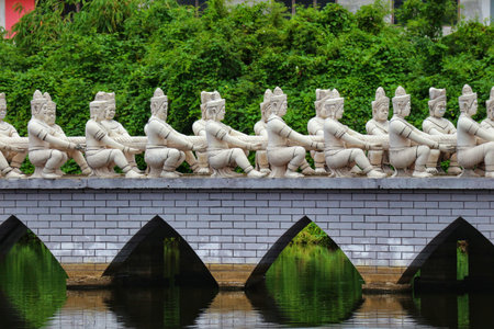 Bangkok, Thailand on 25 July 2023. Row of statues pulling a rope along a bridge over still water with greenery behind, making a tranquil scene, ideal for cultural or travel themed content.の写真素材