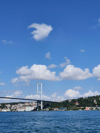 Istanbul, Turkey on 12 July 2022. Bosphorus. A picturesque view of a suspension bridge spanning across the Bosphorus strait in Istanbul. The bridge is prominently featured against a backdrop of a clear blue sky dotted with fluffy white clouds. Lush green hills and coastal buildings line the shoreline, adding to the scenic beauty of the image.の写真素材