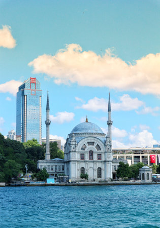Istanbul, Turkey on 12 July 2022. Mosque with minarets sits near a waterway next to a modern city building and lush greenery, creating a scenic cityscape Ideal for travel and cultural themes.のeditorial素材