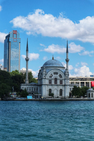 Istanbul, Turkey on 12 July 2022. A beautiful mosque with minarets sits near the water under a blue sky, set against a modern city backdrop, perfect for travel or cultural content.のeditorial素材