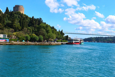 A tranquil river scene showcases a stone tower atop a green hillside and a long suspension bridge under a partly cloudy sky Perfect for travel and architecture photography.のeditorial素材