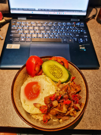 Bekasi, Indonesia 14 Jun 2025. Close-up of a rice bowl with egg, cucumbers, and tomatoes next to a laptop, suggesting meal time during work Perfect for food and technology blogs.のeditorial素材