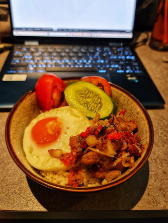 Bekasi, Indonesia 14 Jun 2025. Bowl with rice, fried egg, and vegetables on a desk near a computer provides a quick lunch break option Perfect for illustrating work-life balance.のeditorial素材