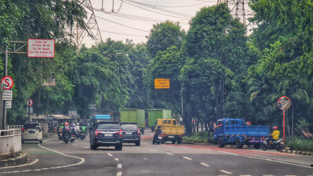 Jakarta, Indonesia on 19 Jun 2025. Various vehicles travel along a city street with lush green trees lining the roadside, creating a busy yet somewhat natural environment Perfect for transportation and urban life concepts.のeditorial素材