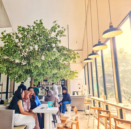 Jakarta, Indonesia on 1 Jun 2025. Group enjoys conversation at bright cafe featuring an indoor tree and ample natural light from large windows Ideal for lifestyle or social gathering content.のeditorial素材