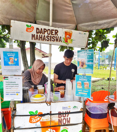 Bogor, Indonesia on 24 Aug 2024. Two people are working at an outdoor herbal tea stand called Dapur Mahasiswa. They are preparing and serving drinks under an umbrella. Several signs advertise the menu and best-selling items. The setting appears to be a paのeditorial素材