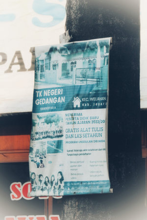 Demak, Indonesia on 5 Oct 2023. A teal and white poster hangs on a post advertising TK Negeri Gedangan Kindergarten in Welahan Jepara. It shows images of the school building and students, and details school supplies and a special program. The poster is slightly faded.の写真素材