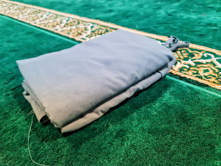 A neatly folded light gray fabric prayer mat rests on a luxurious green carpet with ornate golden border. The simple yet elegant setup suggests a calm and peaceful atmosphere, perfect for prayer or meditation. The soft texture of the fabric and the rich cのeditorial素材