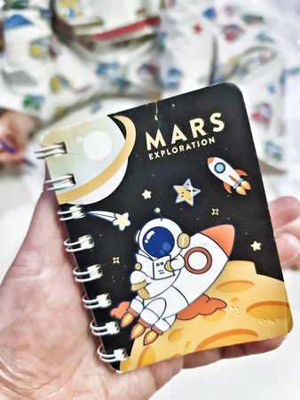 Bekasi, Indonesia on 31 Aug 2024. A small spiral-bound notebook features a charming cartoon astronaut piloting a rocket near a planet. The design incorporates gold accents against a dark background with stars and celestial elements. It's a visually appealのeditorial素材