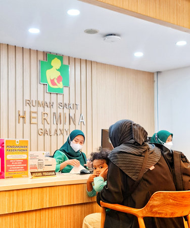 Bekasi, Indonesia on 31 Aug 2024. A family waits at the reception desk of Hermina Galaxy Hospital. A child sits with its mother while a staff member attends to paperwork. The setting is clean and modern, featuring a light wood reception counter and signagのeditorial素材