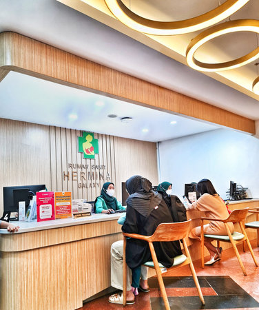 Bekasi, Indonesia on 31 Aug 2024. the reception area of Hermina Galaxy Hospital. Patients in hijabs are sitting at a modern wooden reception desk. The setting is clean, bright, and features circular light fixtures. Informative pamphlets are visible on theのeditorial素材