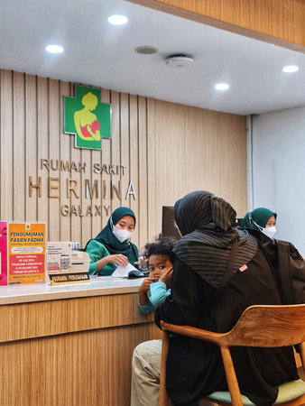 Bekasi, Indonesia on 31 Aug 2024. a family at the reception desk of Hermina Galaxy Hospital. A woman holds a young child while waiting, and hospital staff in masks attend to other patients. The setting is modern and clean, with a wooden reception counter.のeditorial素材