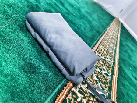 A folded gray musalla prayer mat rests on a green prayer carpet with ornate border. The simple yet elegant design of both the mat and the carpet is highlighted against the soft background. A subtle string detail is visible.のeditorial素材