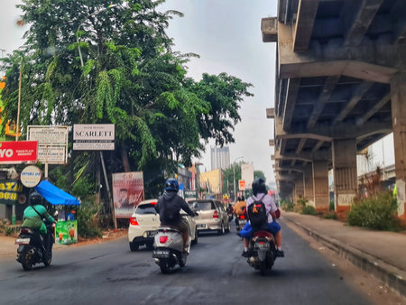 Bekasi, Indonesia on 3 Sep 2024. several motorcyclists traveling under a highway overpass in a city setting. Cars are visible in the background, along with lush green trees and various roadside signs. The overall mood is calm and everyday.のeditorial素材