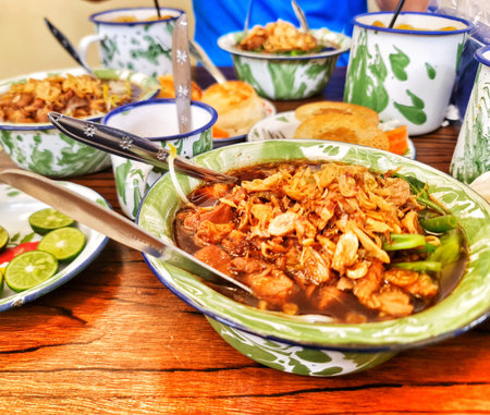Jakarta, Indonesia on 28 Sep 2024. a bowl of Indonesian beef noodle soup garnished with crispy fried onions sits on a wooden table alongside other bowls of food and drinks. The soup is dark brown and rich-looking, served in a vintage-style green and whiteのeditorial素材