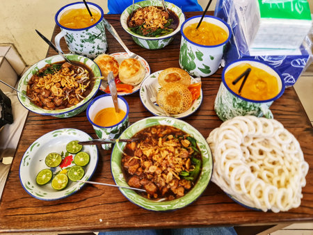 Jakarta, Indonesia on 15 Sep 2024. A table displays various Indonesian dishes in colorful enamel bowls and plates. The food includes noodle soups with fried onions, fried bread, and a unique crispy snack. The setting appears casual, suggesting a local eatのeditorial素材