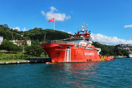 Istanbul, Turkey on 12 July 2022. Orange vessel is docked in a harbor with a flag on a hilltop, under a bright blue sky Perfect for travel and nautical themed projects.のeditorial素材