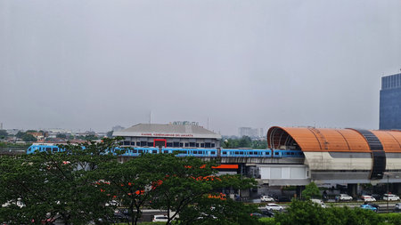 Jakarta, Indonesia on 16 Sept 2025. Blue train moves through urban setting on elevated tracks, viewed from above, featuring green trees, buildings, and overcast skies, suitable for transportation or city life marketing.のeditorial素材