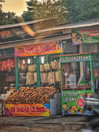 Subang, Indonesia on 4 July 2025. Outdoor food stall displays many brown sweet potatoes and creamy iced beverages, set under a metal awning with banners, creating an authentic street food scene suitable for travel and cultural marketing.のeditorial素材