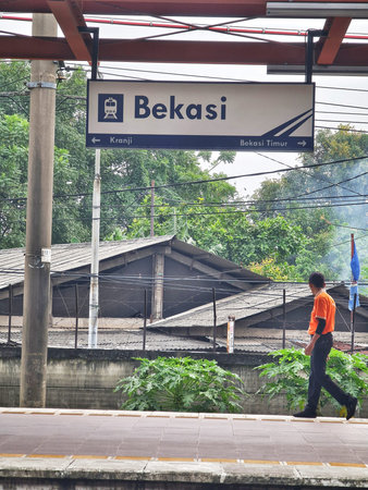 Bekasi, Indonesia on 27 Jun 2025. Man in vibrant orange shirt is walking along gray platform under overhead station signage and surrounding lush green foliage creating an everyday urban scene for lifestyle and travel related content.のeditorial素材