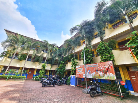 Bekasi, Indonesia on 5 July 2025. Multistory school building shows tropical vibe with lush palm trees along facade, some motorcycles are parked nearby on brick pavement under sunny blue skies; suitable for education or travel content.のeditorial素材