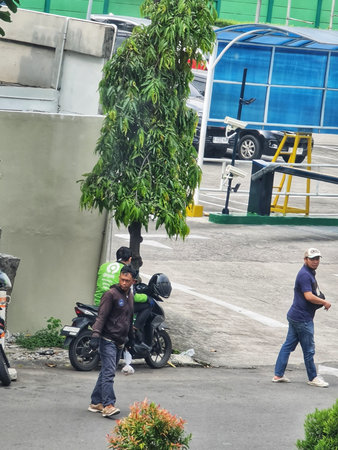 Bekasi, Indonesia on 5 July 2025. Two men walk on an urban street near a motorcycle and a leafy tree, under muted daylight, suggesting a casual everyday scene ideal for lifestyle or travel related visuals.のeditorial素材