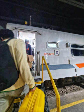 Semarang, Indonesia on 30 Jun 2025. Person with backpack is stepping onto a gray train car at night carrying a vibrant yellow suitcase up metal stairs with yellow railings, suggesting travel and transportation industry imagery.のeditorial素材
