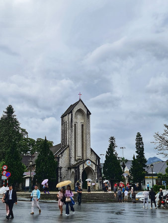 Sa Pa, Vietnam on 15 May 2025. Historic stone church sits amidst a town square on an overcast day, with pedestrians walking and carrying umbrellas, creating a muted and atmospheric scene perfect for travel and architecture-related content.のeditorial素材