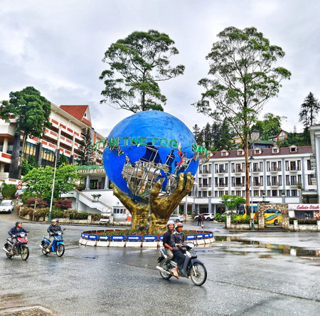 Sa Pa, Vietnam on 15 May 2025. People are riding motorcycles on the wet street near a blue globe sculpture surrounded by buildings and greenery, conveying a cloudy day in an urban setting, ideal for travel and cityscape marketing materials.のeditorial素材