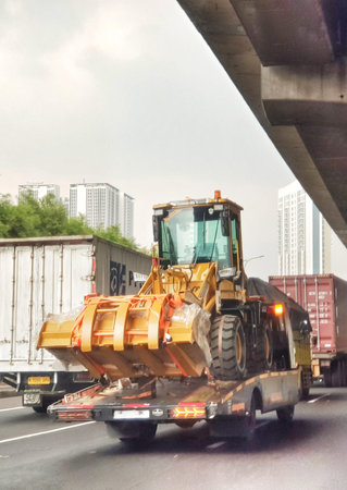 Cikampek, Indonesia on 30 Oct 2025. Yellow front loader sits on a flatbed trailer under an overpass,...