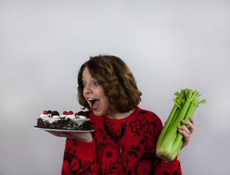 Pretty young woman trying to diet and holding celery and cakeの写真素材