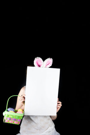 Young woman posing with rabbit ears, Easter basket with sequin eggs and blank sign for copy space on black background の写真素材