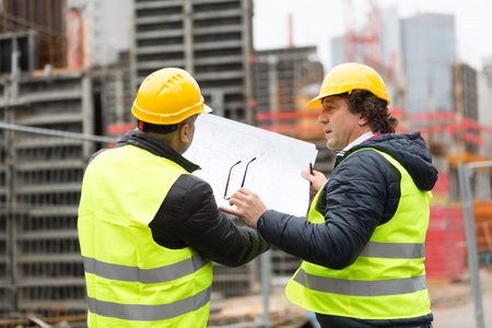 Construction worker with yellow hardhat and safety jacket checking blueprint with an architect