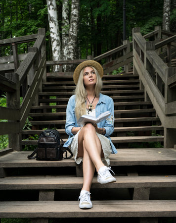 A beautiful young girl with a book in a straw hat is dreamily staring into the sky while sitting on a wooden staircase in the park.の写真素材