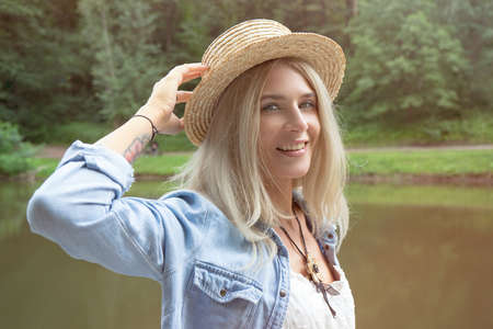 The blonde holds a straw hat on her hand and stands on the edge of a stone pier in a white dress and a blue denim shirt. The concept of vacations, resorts and travel.の写真素材