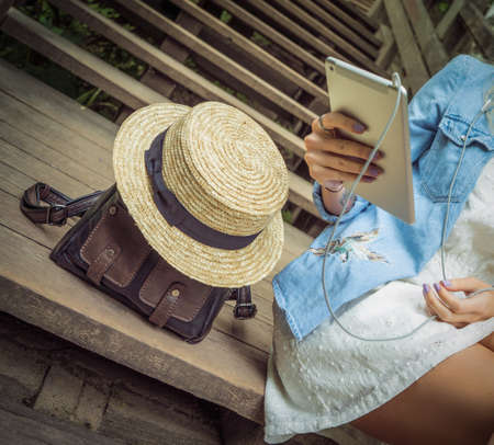 A young girl sits on wooden steps and holds a tablet in her hands, next to her is a leather backpack and a straw hat. The concept of a day off in natureの写真素材