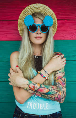 Beautiful young woman hipster with a tattoo in sunglasses and a hat on a bright coloured background. Beach styleの写真素材