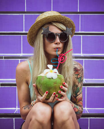 Beautiful young blonde hipster in a straw hat and sunglasses sitting on the purple stairs with coconut in hand. Rain weekend beach partyの写真素材