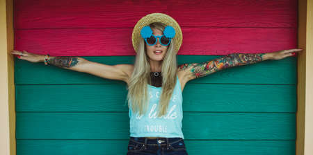 Beautiful young woman hipster with a tattoo in sunglasses and a hat on a bright coloured background. Beach styleの写真素材
