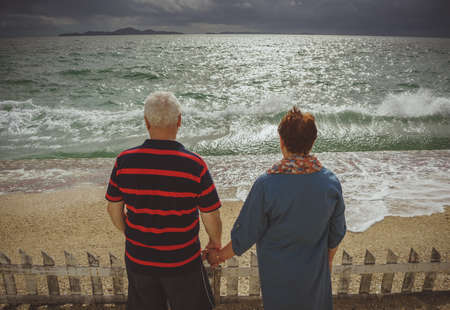 An elderly couple holding hands, looking at the storm at sea. Rear view, back to cameraの写真素材