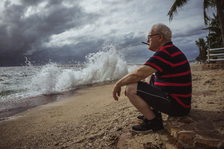 Elderly man in glasses and a striped t-shirt sitting on the beach and looking at the storm smokes a cigar. A wise man in age, the theme of loneliness.の写真素材