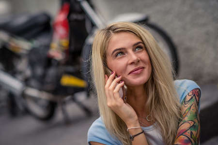 Portrait of young blonde sitting on the background of the motorcycle. Talking on the phone. Female hands with a tattoo. Urban fashion.の写真素材