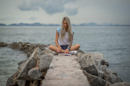 Beautiful blonde woman sitting on a stone embankment and reads book. Hands tattooed. modern, trendy,...