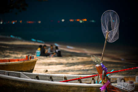 Thai fishing boat with tackle stands on the shore amid vacationers fishermen on the beach.の写真素材